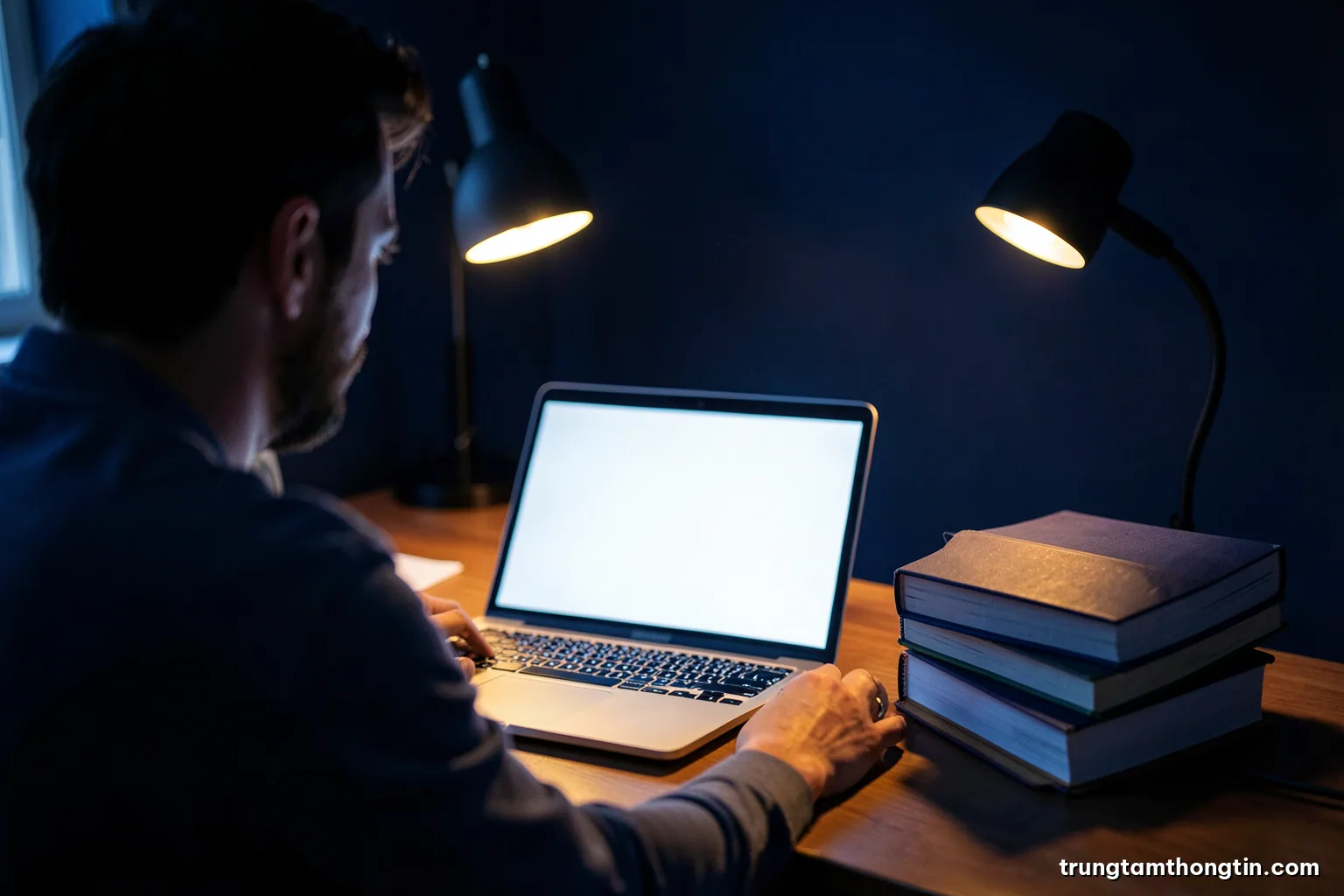 Over-the-shoulder view of a researcher at night comparing a blank glowing laptop with a stack of bound journals, warm desk lamp against deep indigo room, contemplative mood