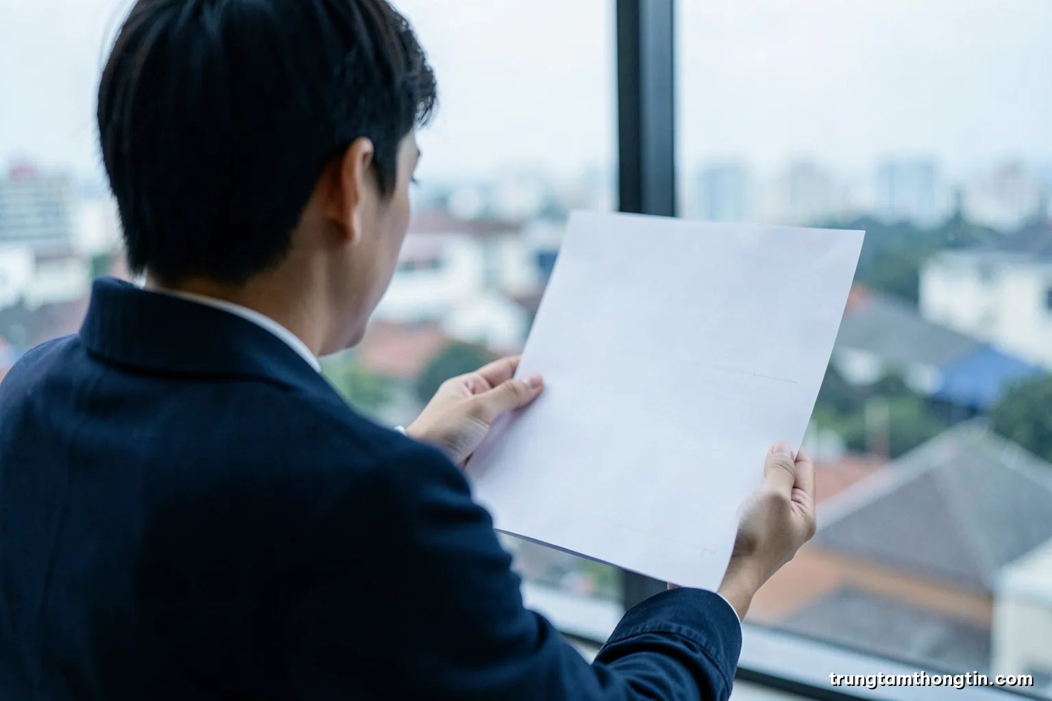 Over-the-shoulder view of a young Vietnamese analyst holding a blank sheet of graph paper up to a window, city rooftops blurred behind, cool overcast light, slate and white palette