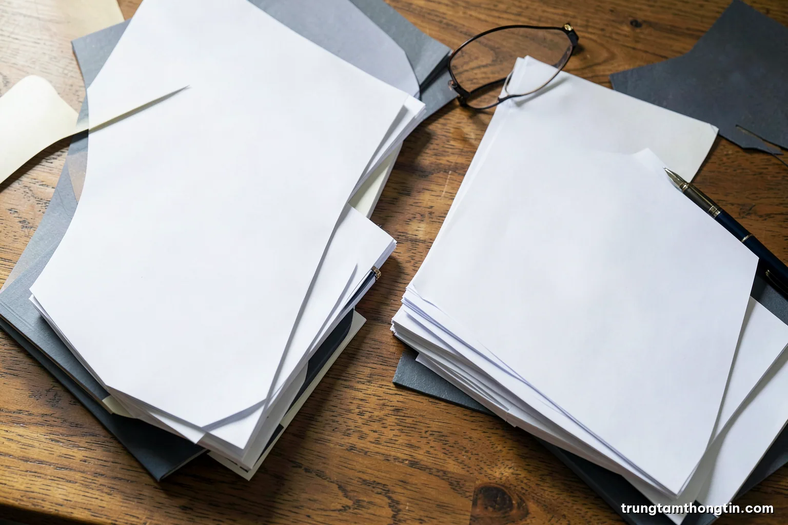Top-down view of a wooden desk covered with stacks of blank white bound reports, a pen and reading glasses, soft north window light, cream and slate grey palette
