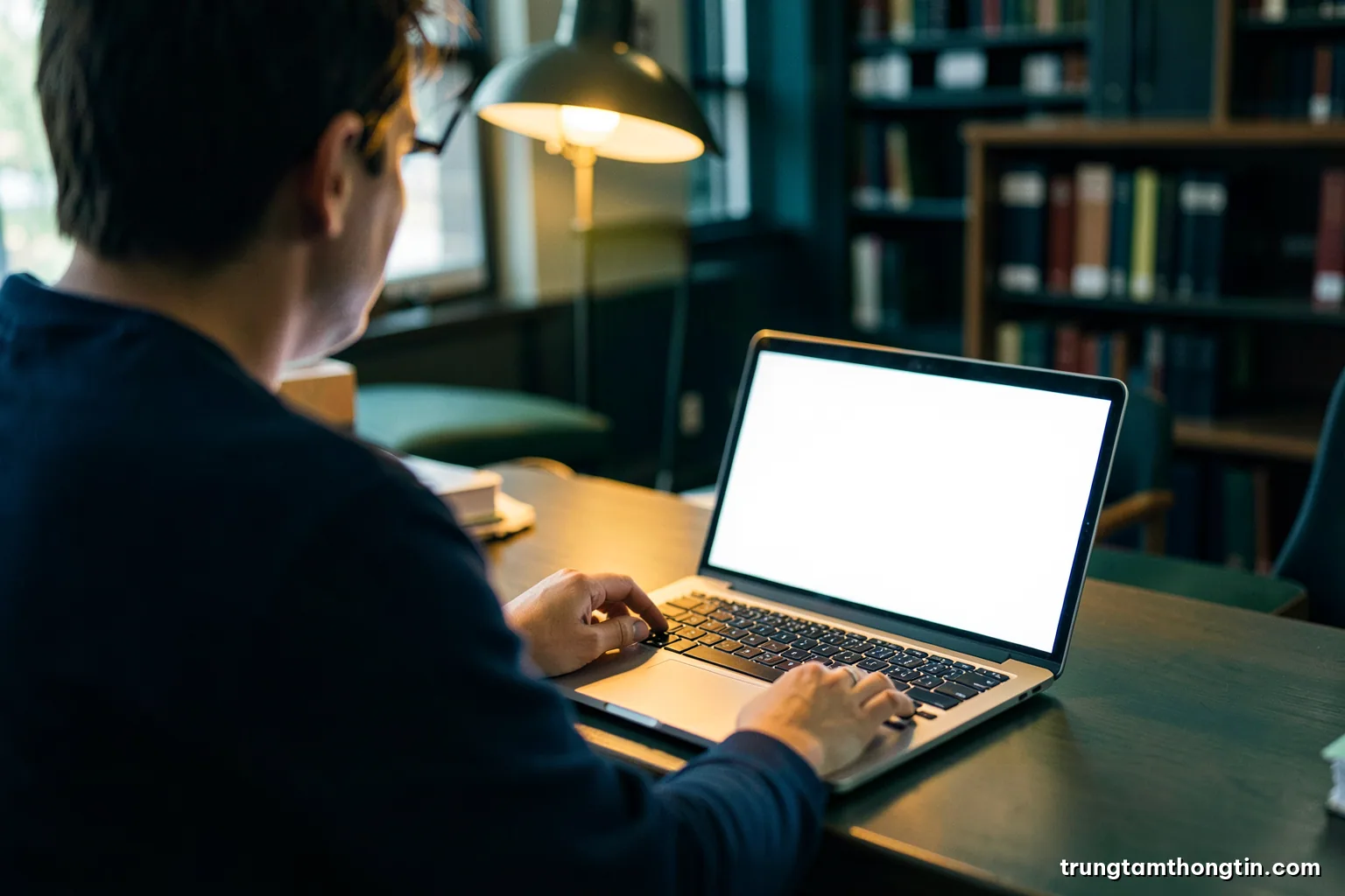 Over-the-shoulder view of a researcher at a university library reading desk with an open laptop showing a blank white display, warm lamp light, deep green palette