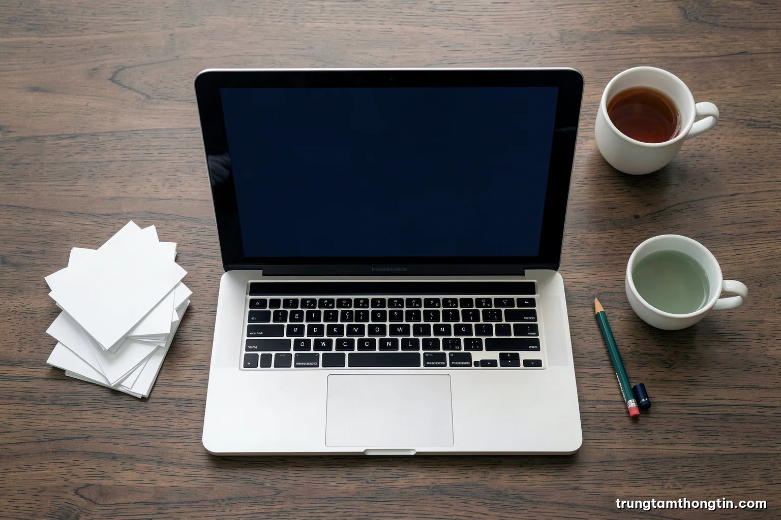 Top-down view of a tidy wooden desk with a laptop showing a blank screen, neatly stacked blank index cards, a pencil and a cup of tea, soft overcast light, sage green palette