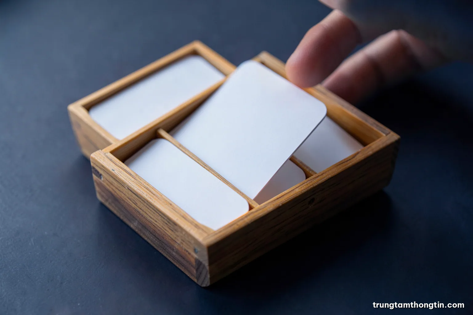 Macro photograph of a small wooden box with five compartments holding blank flashcards, one card held between fingertips, cool north light, slate grey and soft orange palette