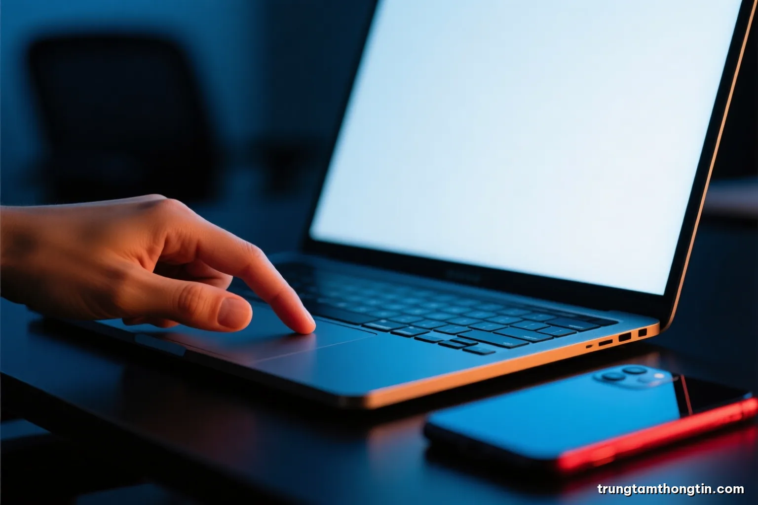 Macro shot of a finger hovering above a laptop trackpad in a dark office, blank glowing screen lighting the hand, a phone face down beside it, cold blue and red rim light