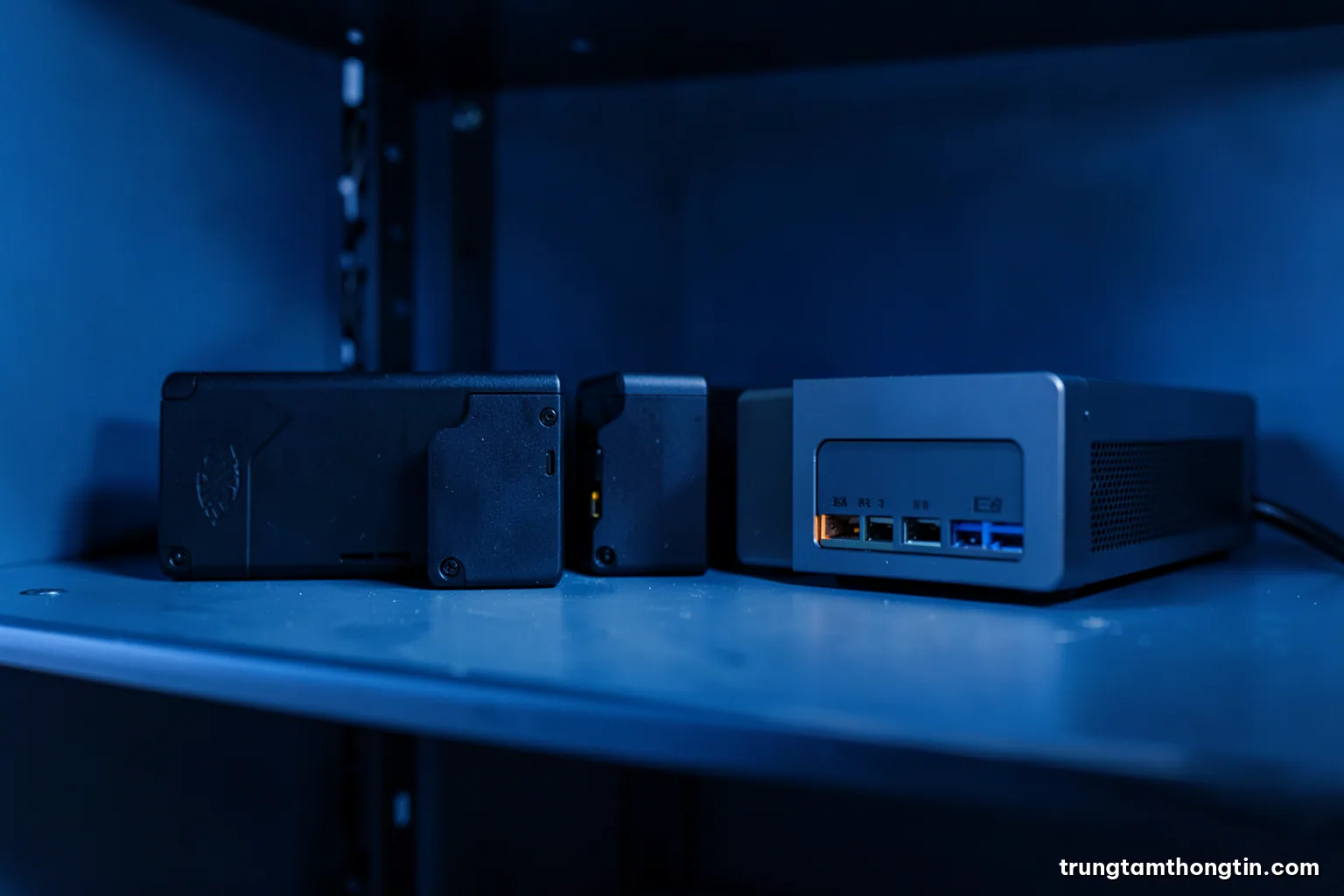 Low angle close-up of three external hard drives and a small network storage box on a metal shelf in a tidy office server corner, cool blue light