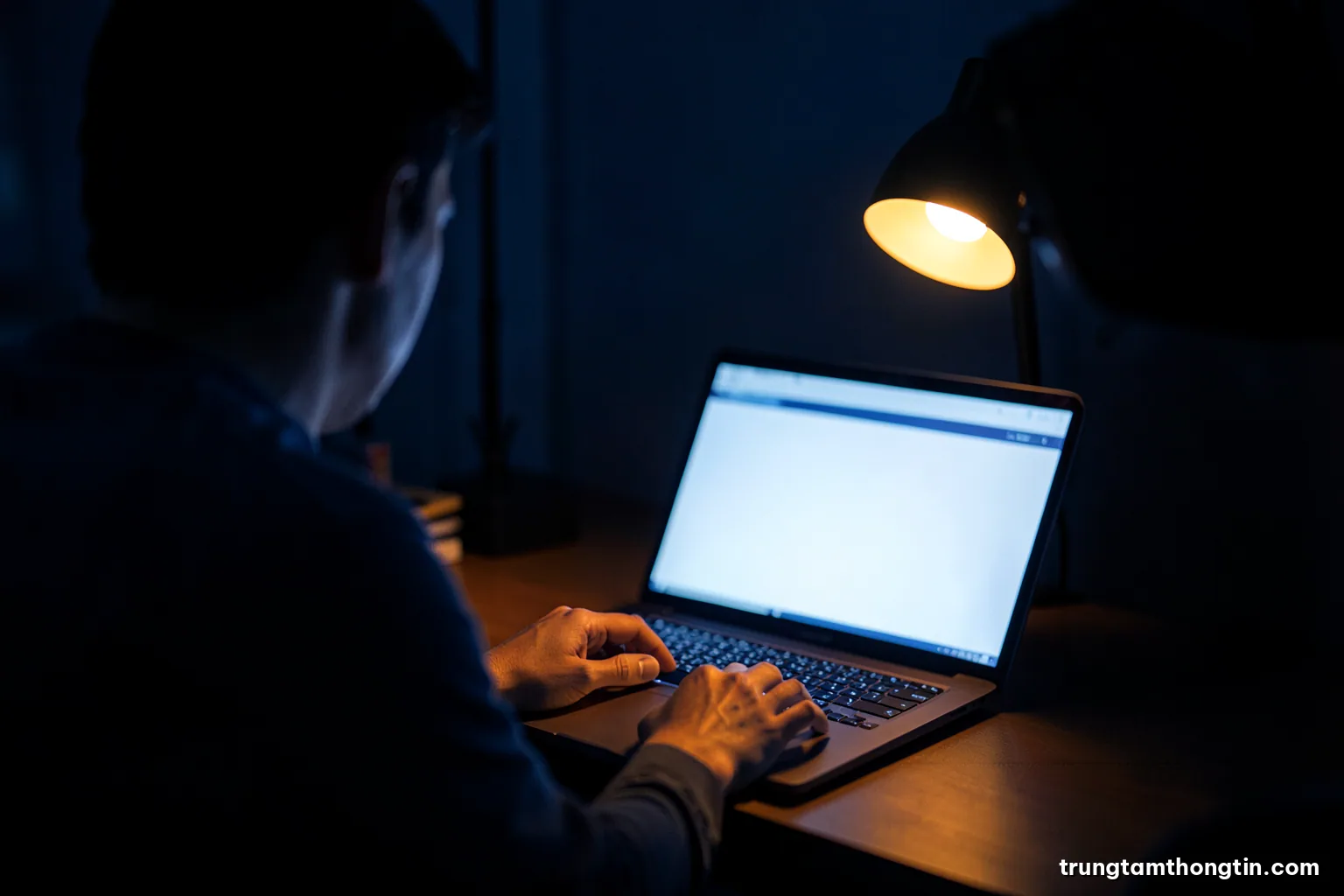 Over-the-shoulder view of a Vietnamese analyst typing on a laptop with a blank glowing display in a dim office at night, desk lamp warm amber, deep navy shadows