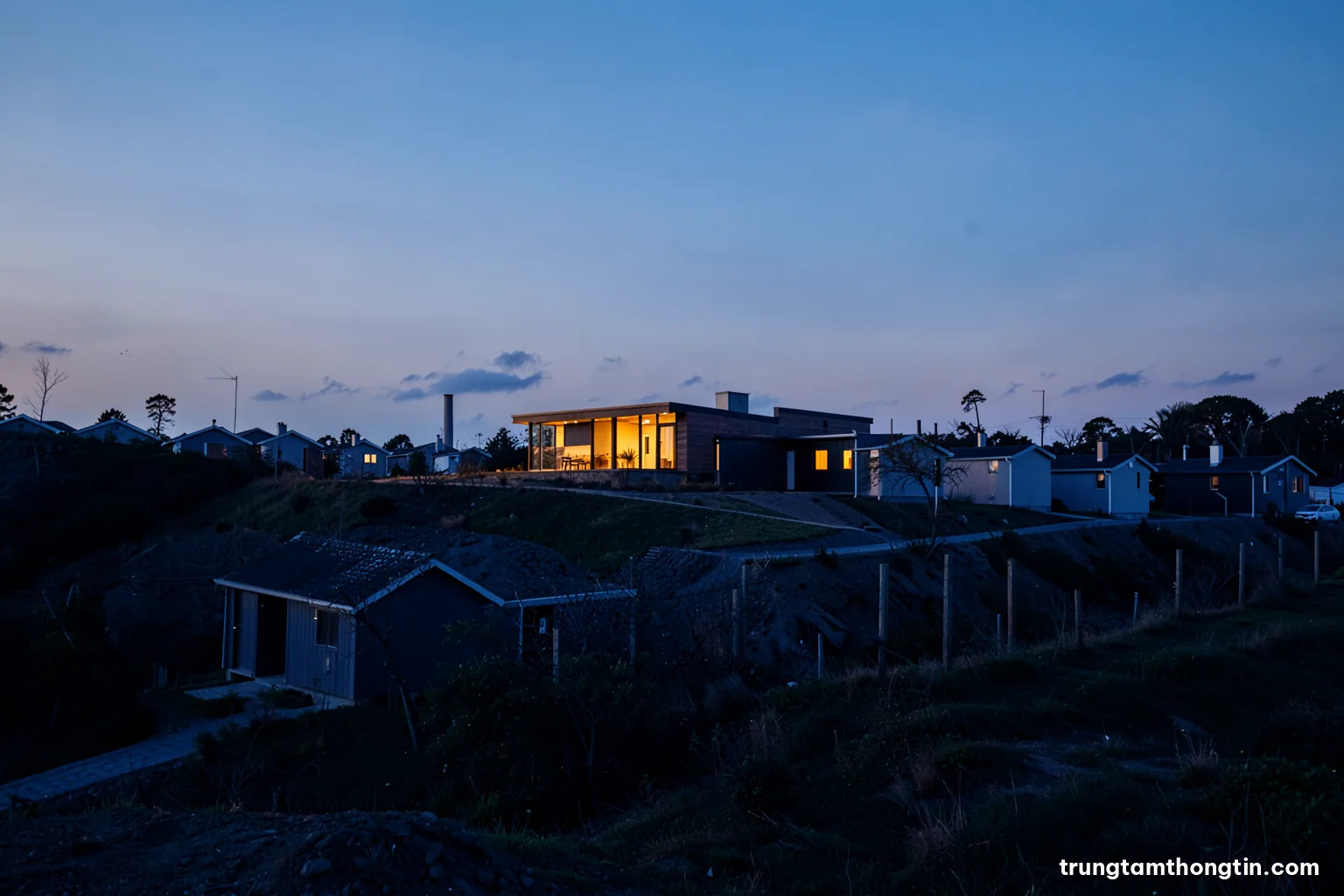 Wide shot of a long row of identical small houses with one large modern villa in the middle, seen from a low hill at dusk, soft blue and amber tones